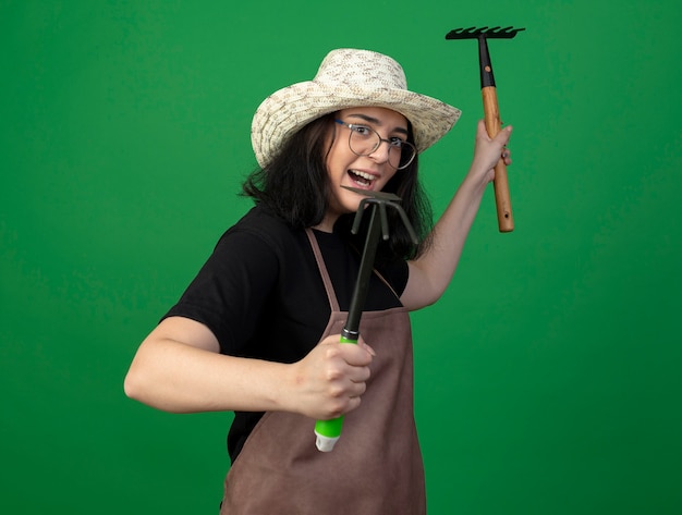 Free photo joyful young brunette female gardener in optical glasses and uniform wearing gardening hat stands sideways holding rake and hoe rake isolated on green wall