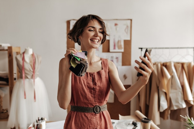 Joyful young brunette short-haired woman in linen red dress smiles sincerely, holds pieces of textiles and takes selfie in office