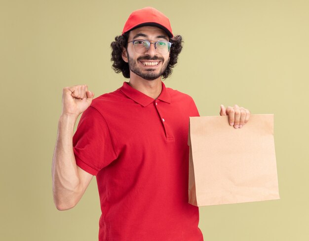 Joyful young caucasian delivery man in red uniform and cap wearing glasses holding paper package doing be strong gesture 