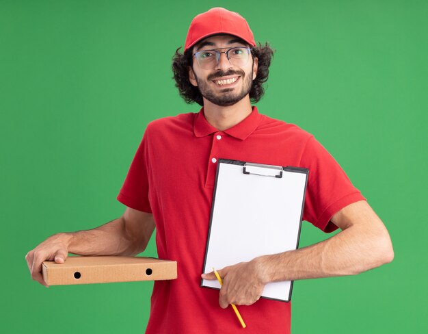 Joyful young caucasian delivery man in red uniform and cap wearing glasses holding pizza package pencil showing clipboard to camera 