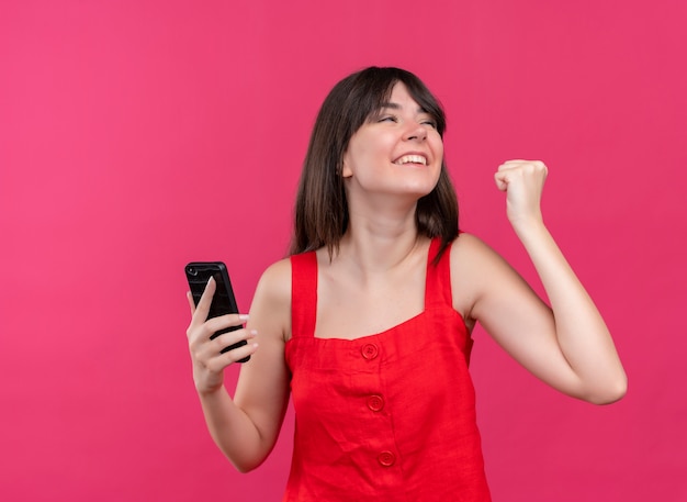 Joyful young caucasian girl holding phone and holding fist up looking to the side on isolated pink background with copy space