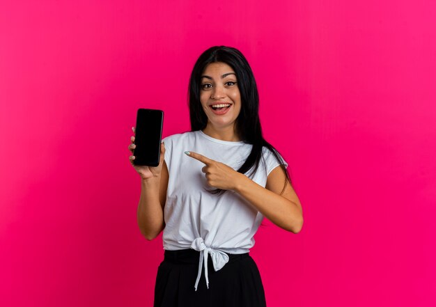 Joyful young caucasian woman holds and points at phone