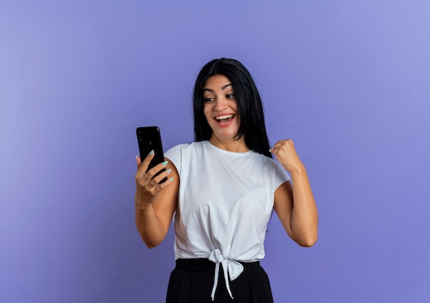 Joyful young caucasian woman keeps fist and looks at phone