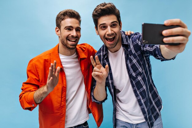 Joyful young charming men take selfie in great mood on blue background Cool man in orange jacket waves hand in greeting Guy in checkered shirt holds phone and shows peace sign