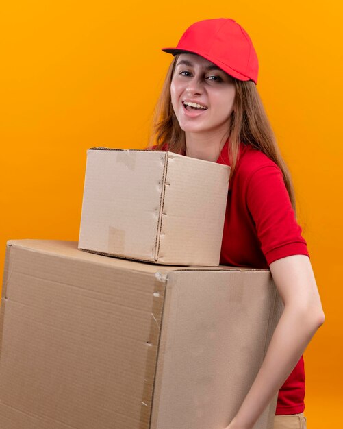 Joyful young delivery girl in red uniform holding boxes on isolated orange space