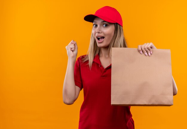 Joyful young delivery woman wearing red uniform and cap holding paper bag and showing yes gesture 