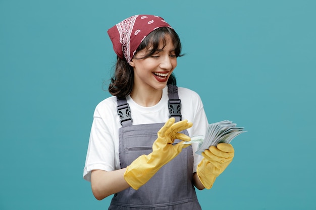 Free photo joyful young female cleaner wearing uniform bandana and rubber gloves holding money looking at it counting money isolated on blue background