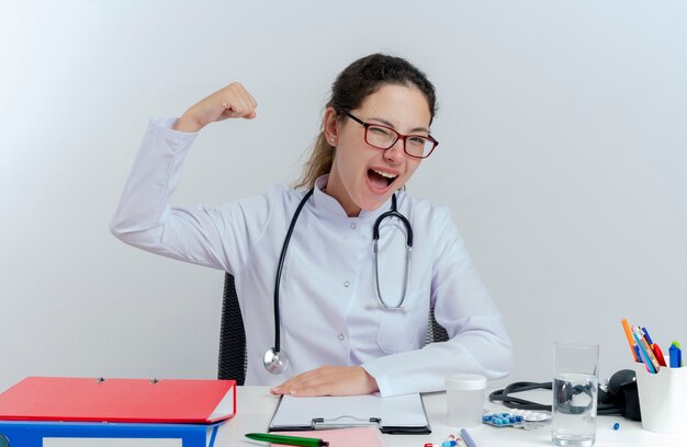 Joyful young female doctor wearing medical robe and stethoscope and glasses sitting at desk with medical tools looking winking doing strong gesture isolated