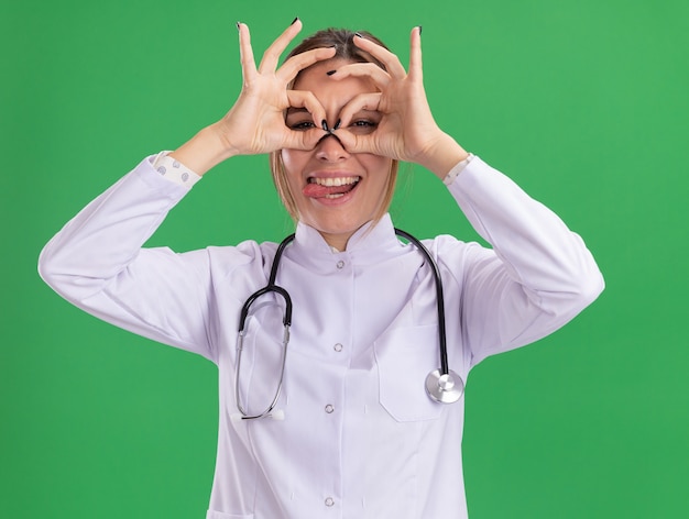 Joyful young female doctor wearing medical robe with stethoscope showing look gesture isolated on green wall