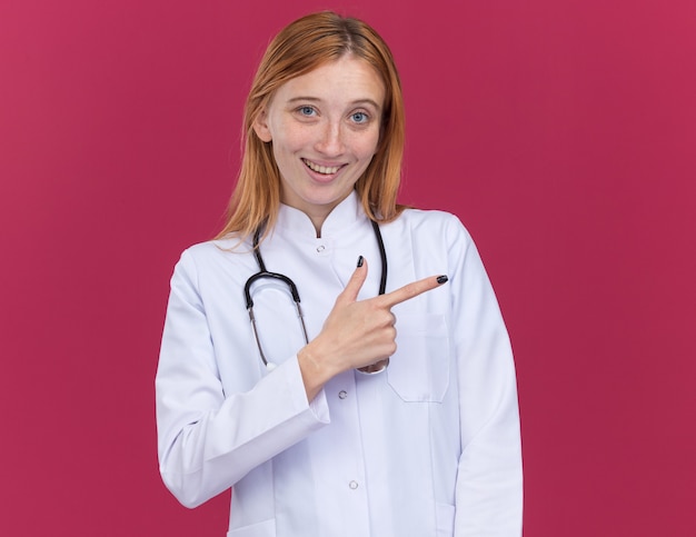 Joyful young female ginger doctor wearing medical robe and stethoscope looking at front pointing at side isolated on crimson wall with copy space