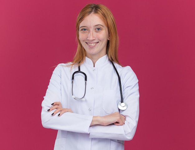 Free photo joyful young female ginger doctor wearing medical robe and stethoscope standing with closed posture  isolated on crimson wall with copy space