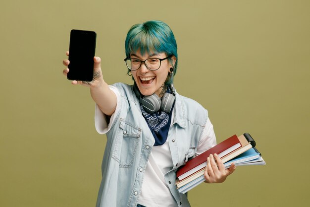 Joyful young female student wearing glasses bandana and headphones around neck holding note pads looking at camera stretching mobile phone out towards camera isolated on olive green background