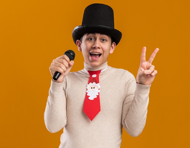 Joyful young guy wearing hat with christmas tie speaks on microphone showing peace gesture isolated on yellow wall