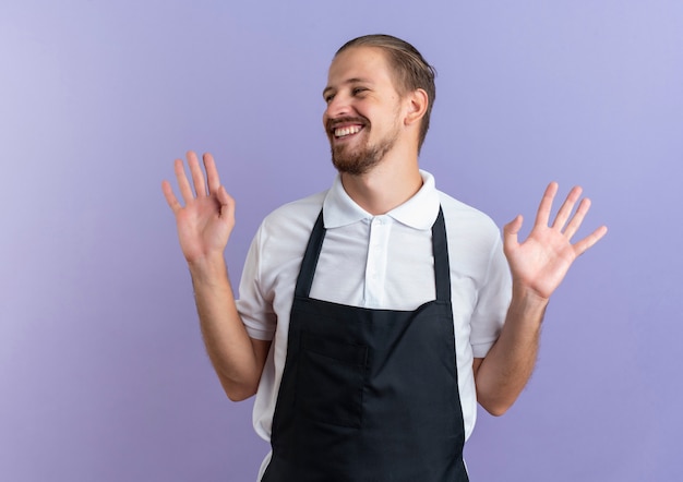 Free photo joyful young handsome barber wearing uniform showing empty hands and looking at side isolated on purple