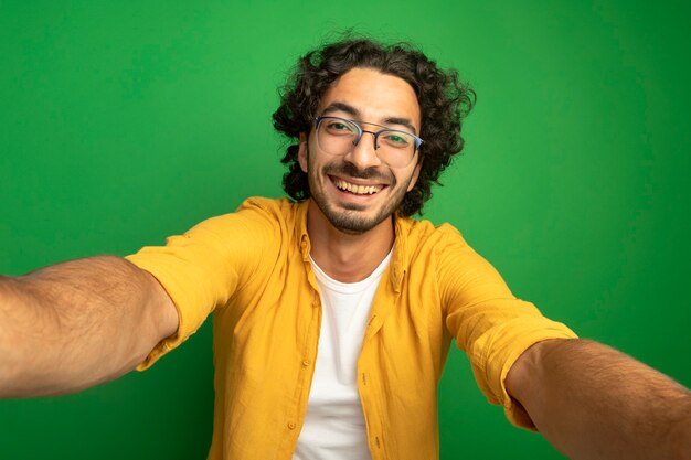 Joyful young handsome caucasian man wearing glasses looking at camera stretching out hands towards camera isolated on green background