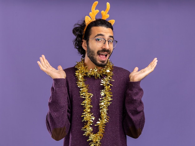 Free Photo joyful young handsome guy wearing christmas hair hoop with garland on neck spreading hands isolated on blue wall