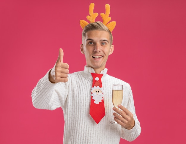 Joyful young handsome guy wearing reindeer antlers headband and santa claus tie holding glass of champagne  showing thumb up isolated on pink wall with copy space