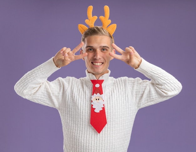 Joyful young handsome guy wearing reindeer antlers headband and santa claus tie looking at camera showing v-sign symbols near eyes isolated on purple background