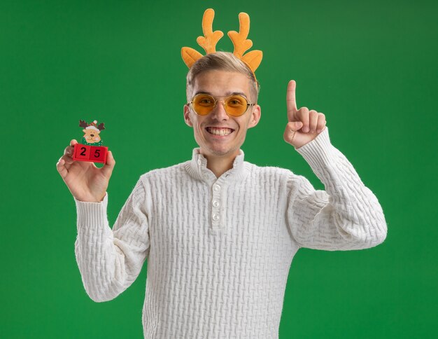 Joyful young handsome guy wearing reindeer antlers headband with glasses holding christmas tree toy with date looking at camera pointing up isolated on green background