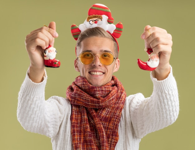 Joyful young handsome guy wearing santa claus headband and scarf  stretching out santa claus christmas ornaments towards isolated on olive green wall