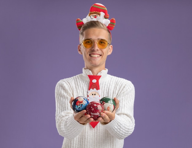 Free photo joyful young handsome guy wearing santa claus headband and tie with glasses holding christmas ball ornaments looking at camera isolated on purple background