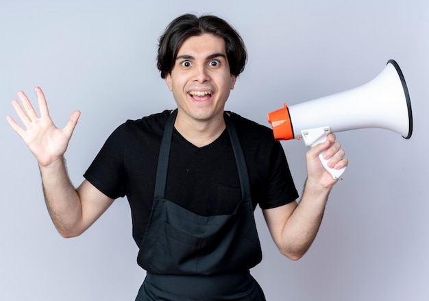 Joyful young handsome male barber in uniform holding loudspeaker and spread hand isolated on white background