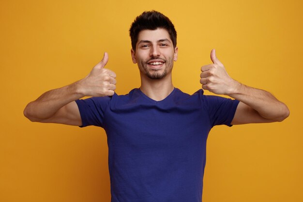 Joyful young handsome man looking at camera showing thumbs up on yellow background