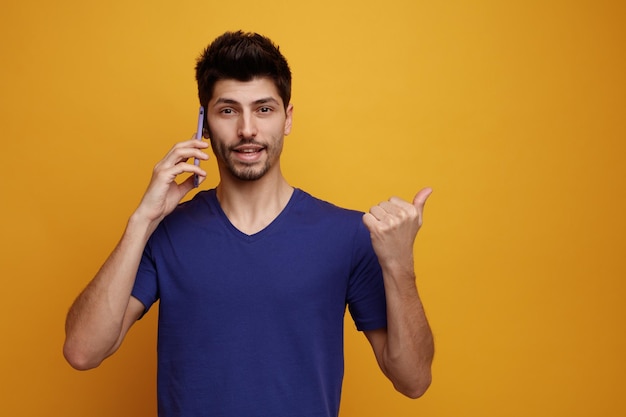 Joyful young handsome man talking on phone looking at camera pointing to side on yellow background