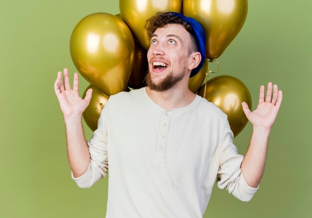 Joyful young handsome slavic party guy wearing party hat standing in front of balloons showing empty hands looking up isolated on olive green background