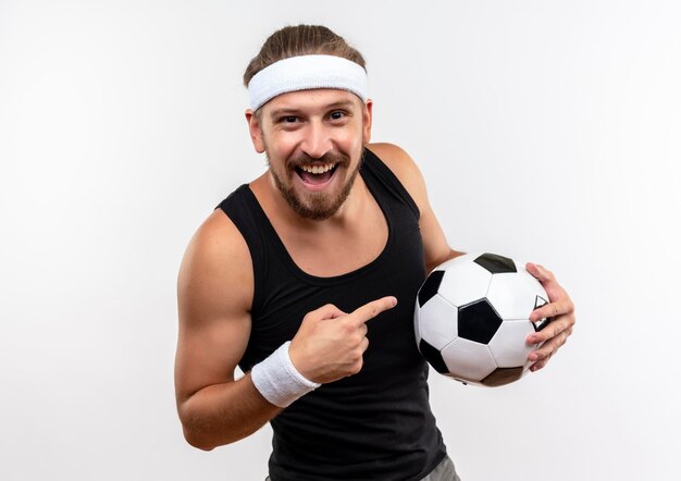 Joyful young handsome sporty man wearing headband and wristbands holding and pointing at soccer ball isolated on white wall