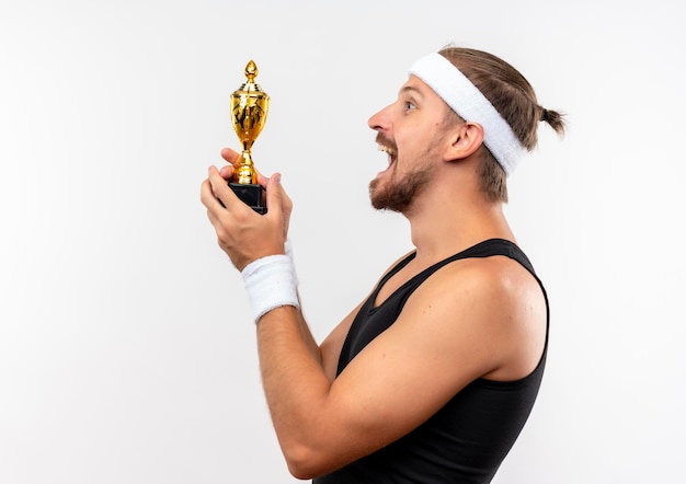 Free photo joyful young handsome sporty man wearing headband and wristbands standing in profile view holding winner cup isolated on white wall with copy space