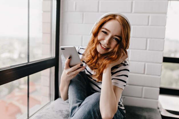 Free photo joyful young lady in headphones enjoying weekend. attractive ginger girl with phone sitting beside window.