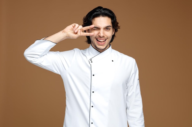 Free photo joyful young male chef wearing uniform looking at camera showing peace sign near eye winking isolated on brown background