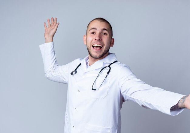 Joyful young male doctor wearing medical robe and stethoscope around his neck stretching out arm and raising hand isolated on white