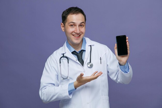 Joyful young male doctor wearing medical robe and stethoscope around neck looking at camera showing mobile phone pointing hand at it isolated on purple background