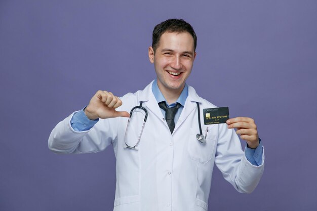 Joyful young male doctor wearing medical robe and stethoscope around neck showing credit card pointing at it looking at camera isolated on purple background