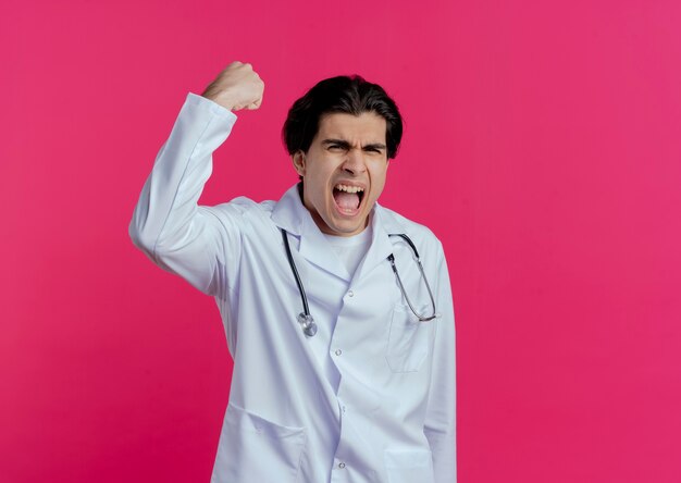 Joyful young male doctor wearing medical robe and stethoscope  doing yes gesture isolated on pink wall with copy space