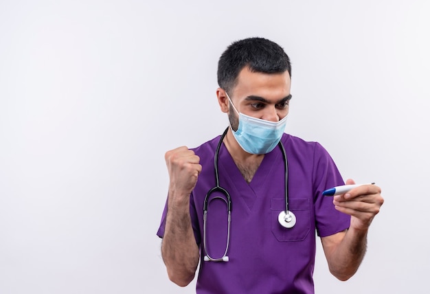 Joyful young male doctor wearing purple surgeon clothing and stethoscope medical mask looking at thermometer in his hand showing yes gesture on isolated white wall