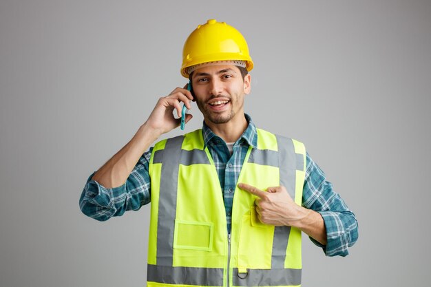 Joyful young male engineer wearing safety helmet and uniform looking at camera pointing at himself while talking on phone isolated on white background