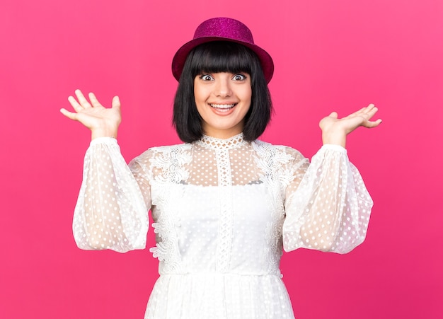 Joyful young party girl wearing party hat  showing empty hands isolated on pink wall