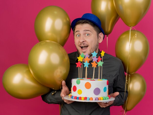 Joyful young party guy wearing blue hat standing among balloons holding cake isolated on pink