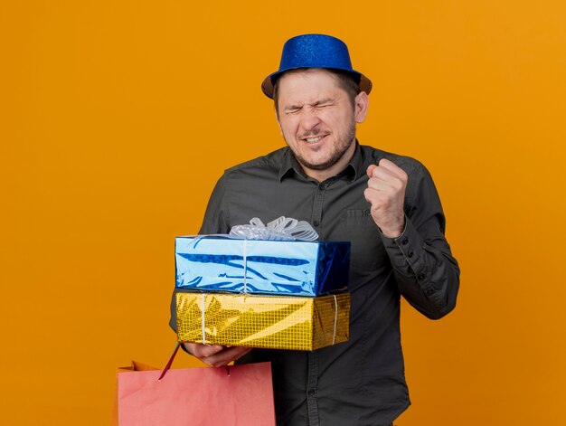 Joyful young party guy with closed eyes wearing blue hat holding gift boxes and bag showing yes gesture isolated on orange