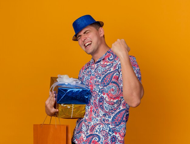 Joyful young party guy with closed eyes wearing blue hat holding gift boxes with bag showing yes gesture isolated on orange