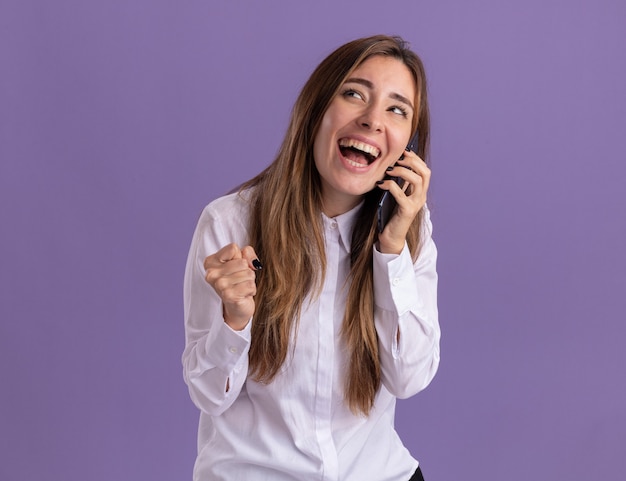 Joyful young pretty caucasian girl keeps fist and talks on phone looking at side on purple 