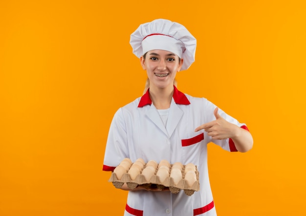 Joyful young pretty cook in chef uniform with dental braces holding and pointing at carton of eggs isolated on orange space 