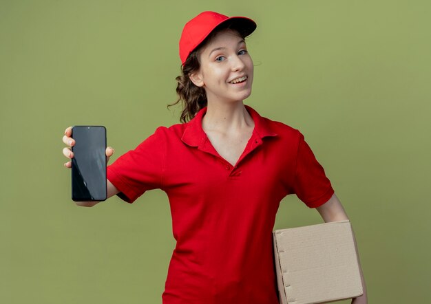 Joyful young pretty delivery girl in red uniform and cap holding carton box and stretching out mobile phone at camera isolated on olive green background