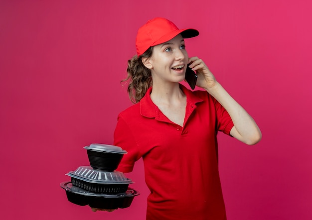 Free photo joyful young pretty delivery girl wearing red uniform and cap talking on phone and holding food containers looking at side  on crimson space