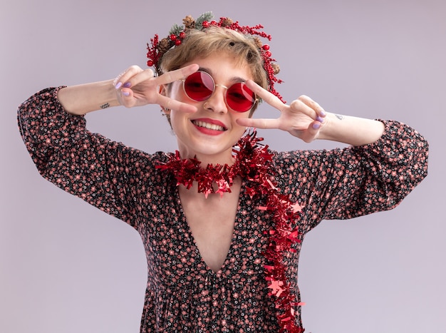 Free photo joyful young pretty girl wearing christmas head wreath and tinsel garland around neck with glasses looking at camera showing v-sign symbols near eyes isolated on white background