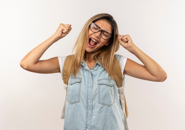 Joyful young pretty student girl wearing glasses and back bag raising fists with closed eyes isolated on white