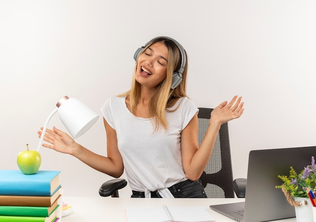 Joyful young pretty student girl wearing headphones sitting at desk with school tools listening to music showing empty hands with closed eyes isolated on white